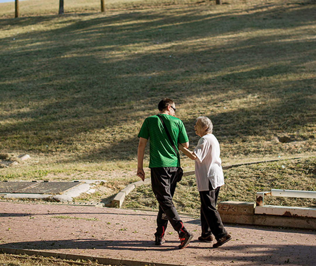 Eine ältere Dame hakt sich bei einem jungen Mann unter. Gemeinsam gehen sie in einem Park spazieren und unterhalten sich