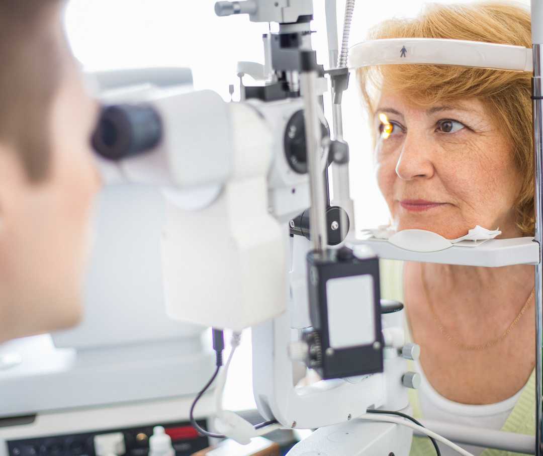 Das Foto zeigt einen Augenarzt, der mithilfe eines Untersuchungsgeräts die Augen einer älteren Frau betrachtet. Der Arzt ist links im Bild im Anschnitt und unscharf zu sehen. Er blickt mit seinen Augen in das Untersuchungsgerät. Die Frau ist rechts im Bild scharf zu sehen. Sie blickt von der anderen Seite ins Untersuchungsgerät. Ein Lichtstrahl ist auf ihrem rechten Auge zu sehen. Sie hat halblange rötliche Haare und trägt einen mintfarbenen Pullover und ein weißes T-Shirt.
