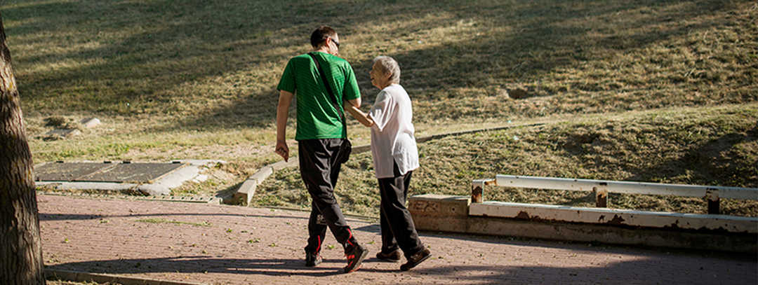 Ratgeber Makula: Eine ältere Dame hakt sich bei einem jungen Mann unter. Eine ältere Dame hakt sich bei einem jungen Mann unter. Gemeinsam gehen sie in einem Park spazieren und unterhalten sich.