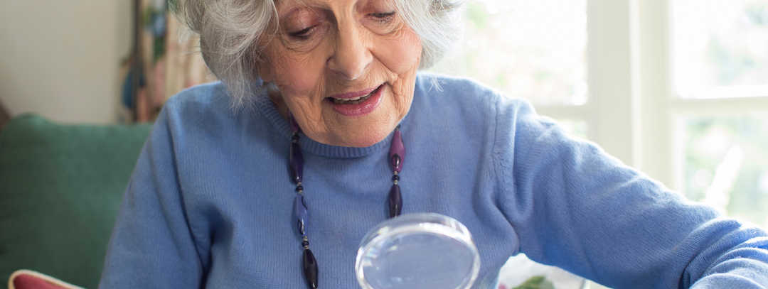 Eine gut gelaunte ältere Dame liest mithilfe einer Lupe in einem Buch.