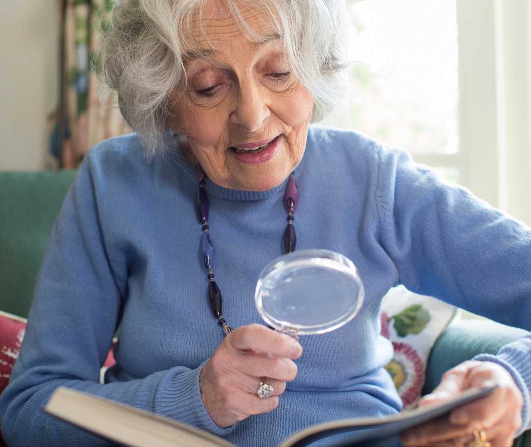 Eine gut gelaunte ältere Dame liest mithilfe einer Lupe in einem Buch