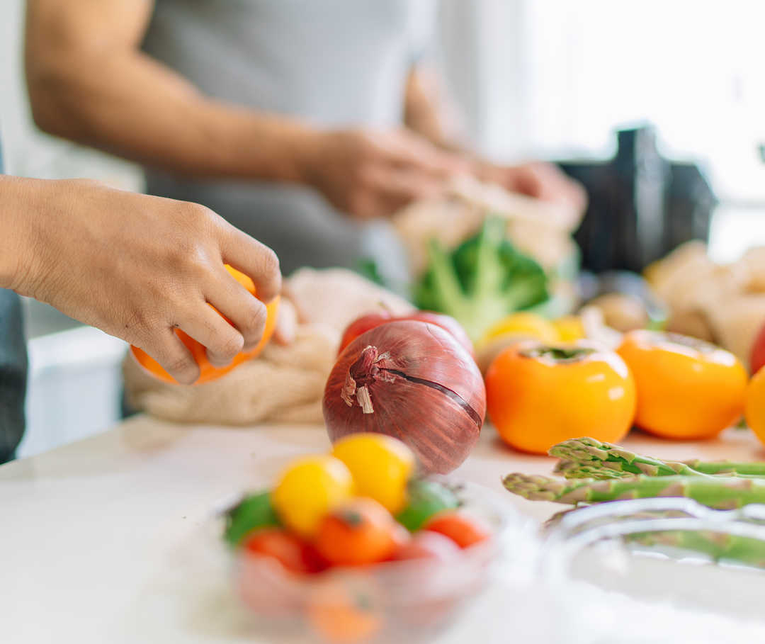 Das Bild zeigt verschiedene Obst- und Gemüsesorten, die auf einer Arbeitsfläche liegen. In den Bildausschnitt ragen die Hände von zwei Personen, die das Obst und Gemüse waschen und klein schneiden.