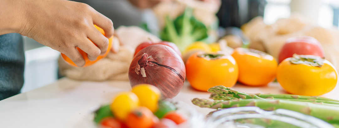 Diabetische Netzhauterkrankungen: Ernährung Das Bild zeigt verschiedene Obst- und Gemüsesorten, die auf einer Arbeitsfläche liegen. In den Bildausschnitt ragen die Hände von zwei Personen, die das Obst und Gemüse waschen und klein schneiden.